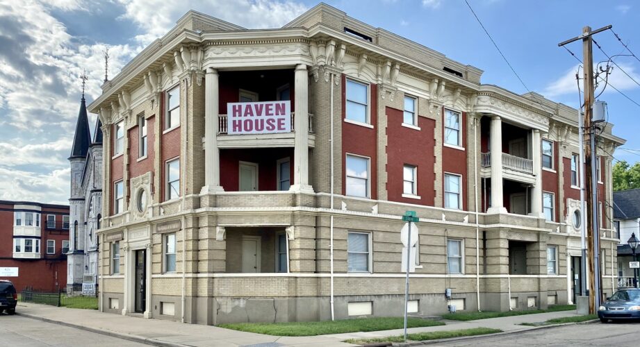 A large, three-story brick building on a street corner, featuring tan stone accents and tall columns. A balcony on the second floor displays a large sign that reads “Haven House” in red letters. The sky is partly cloudy, and nearby buildings and a church with a steeple are visible in the background.