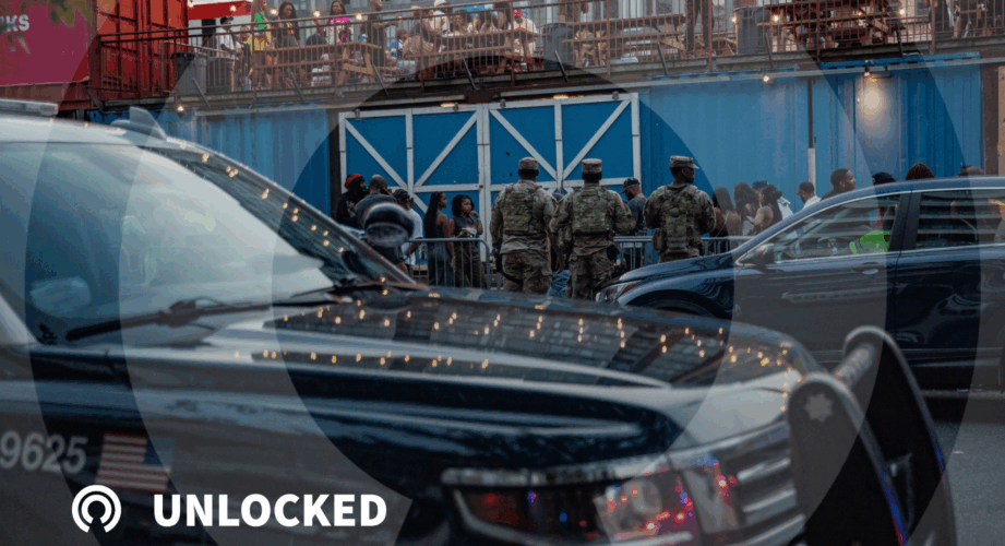 A group of military personnel in camouflage uniforms stand on a Washington, D.C., street near a crowd of people, with a police car in the foreground. In the background, people are seated on an elevated outdoor patio decorated with string lights, and blue shipping containers form part of the scene.