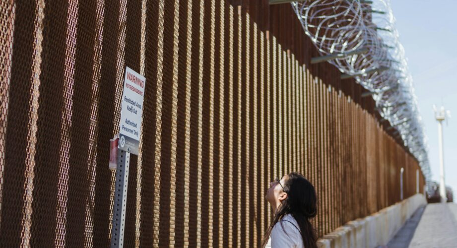 Woman standing at the base of a tall metal border fence topped with coiled razor wire, looking up near a “Warning: No Trespassing, Restricted Area, Keep Out, Authorized Personnel Only” sign.