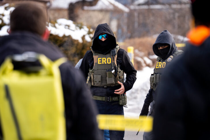 Two masked law enforcement officers in heavy tactical gear with “ERO” on their vests stand outdoors in a snowy residential area, partially framed by blurred figures and yellow caution tape in the foreground.