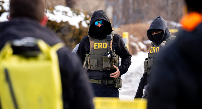 Two masked law enforcement officers in heavy tactical gear with “ERO” on their vests stand outdoors in a snowy residential area, partially framed by blurred figures and yellow caution tape in the foreground.
