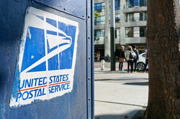 Close-up of a weathered blue United States Postal Service mailbox with the USPS eagle logo, on a city sidewalk with a tree, buildings, and pedestrians blurred in the background.