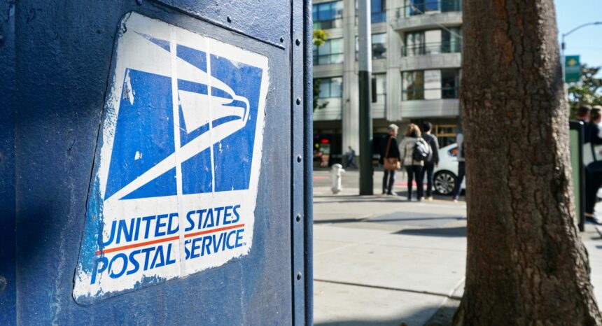 Close-up of a weathered blue United States Postal Service mailbox with the USPS eagle logo, on a city sidewalk with a tree, buildings, and pedestrians blurred in the background.
