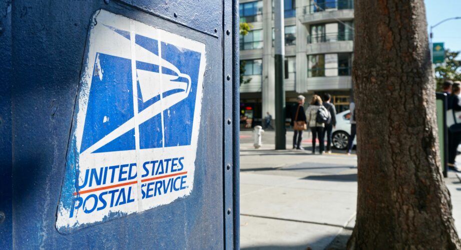 Close-up of a weathered blue United States Postal Service mailbox with the USPS eagle logo, on a city sidewalk with a tree, buildings, and pedestrians blurred in the background.
