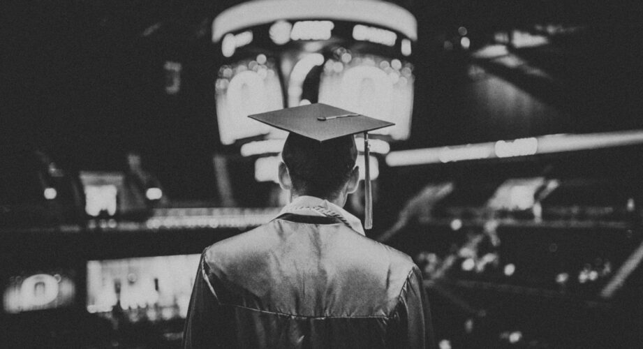 Black-and-white photo of a graduate seen from behind, wearing a cap and gown, standing in a large indoor arena with bright, out-of-focus screens and lights in the background.