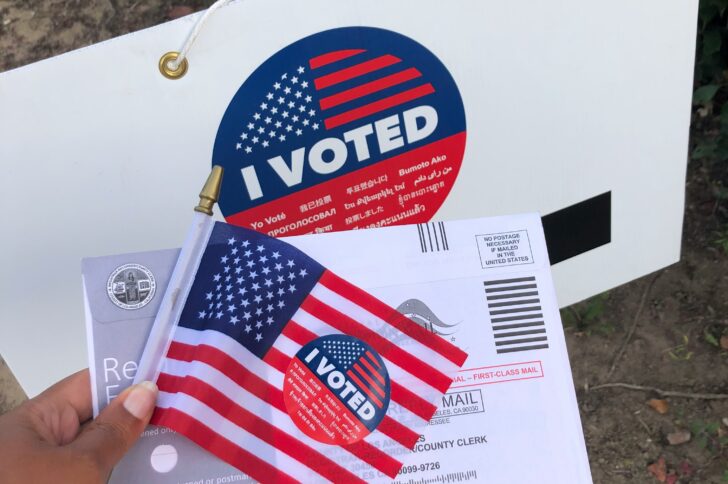 A hand holds a small American flag and a mail‑in ballot envelope, both with round red, white, and blue “I VOTED” stickers, in front of a white sign that also displays a large “I VOTED” sticker.