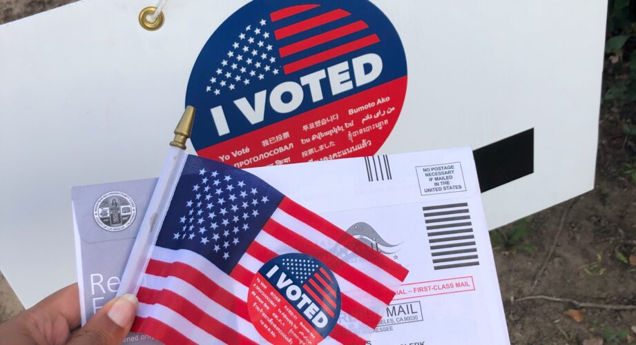 A hand holds a small American flag and a mail‑in ballot envelope, both with round red, white, and blue “I VOTED” stickers, in front of a white sign that also displays a large “I VOTED” sticker.