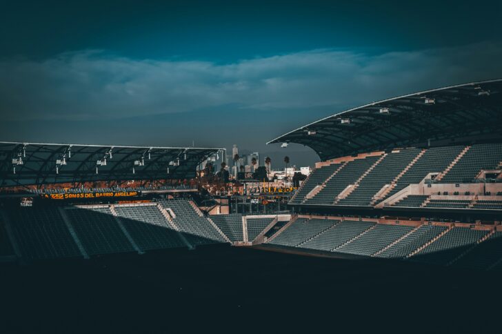 Wide-angle view of an empty outdoor sports stadium at dusk, rows of gray seats in shadow, two covered stands framing the scene, with palm trees and a distant city skyline under a dark blue, cloudy sky.