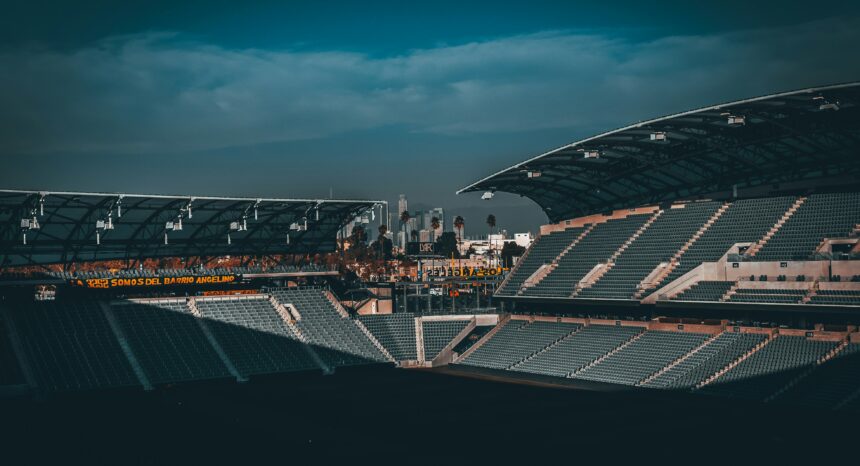 Wide-angle view of an empty outdoor sports stadium at dusk, rows of gray seats in shadow, two covered stands framing the scene, with palm trees and a distant city skyline under a dark blue, cloudy sky.