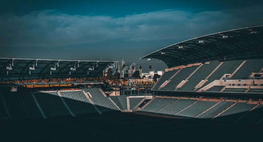 Wide-angle view of an empty outdoor sports stadium at dusk, rows of gray seats in shadow, two covered stands framing the scene, with palm trees and a distant city skyline under a dark blue, cloudy sky.